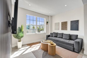 A modern living room with a grey sofa, a wooden coffee table, a potted plant, and a large window. - Cozy 1BR in the Heart of SoBe Ocean 1 Min Away 305 (Miami Beach)