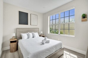 Une chambre avec une grande fenêtre, un lit recouvert de draps blancs, deux tables de chevet et une petite plante accrochée au mur.
