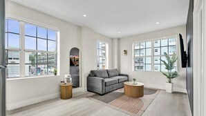 Un salón amplio con ventanas grandes, un sofá gris, dos mesitas redondas y un televisor de pantalla plana montado en la pared.