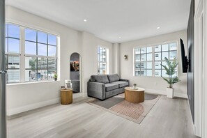 A spacious living room with large windows, a grey sofa, two round side tables, and a flat-screen TV mounted on the wall. - Prime South Beach 1BR/1BA Beach 1 Min Away 301 (Miami Beach)