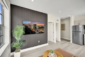 A modern living room with a flat-screen TV mounted on a dark, textured wall, a wooden coffee table, a potted plant, and a stainless steel refrigerator. - Prime South Beach 1BR/1BA Beach 1 Min Away 301 (Miami Beach)