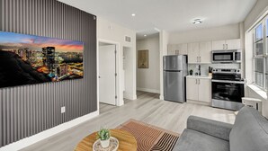 Ein modernes Wohnzimmer mit Flachbildfernseher, grauem Sofa, Couchtisch aus Holz und einer Küche mit Edelstahlgeräten.