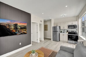 A modern living room with a flat-screen TV, a grey sofa, a wooden coffee table, and a kitchen with stainless steel appliances. - Modern 1BR South Beach Gem 1 Min Walk to Beach 201 (Miami Beach)