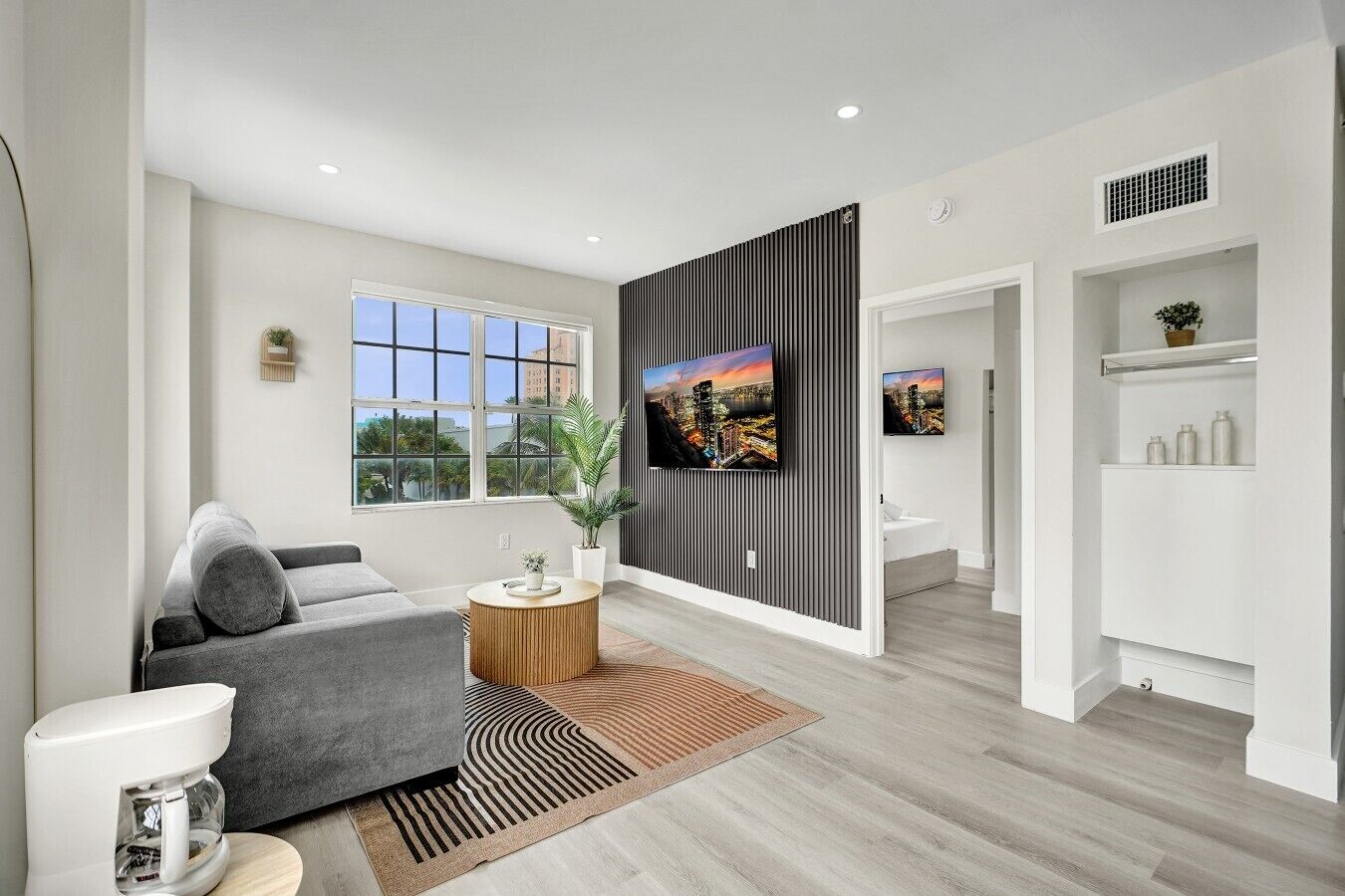 A modern living room with a grey sofa, a coffee machine, a TV mounted on a wall, and a large window with a city view.