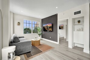 A modern living room with a grey sofa, a coffee machine, a TV mounted on a wall, and a large window with a city view. - Modern 1BR South Beach Gem 1 Min Walk to Beach 201 (Miami Beach)