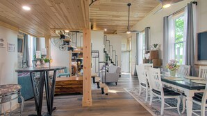 Une pièce spacieuse avec un plafond en bois, un coin repas avec des chaises blanches, une grande fenêtre avec des rideaux et un ventilateur de plafond.