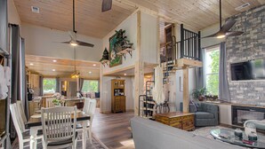 Un vaste espace de vie doté d’un haut plafond, de poutres en bois, d’une cheminée, d’un téléviseur fixé à un mur en pierre, ainsi que d’un coin repas avec une table et des chaises en bois.