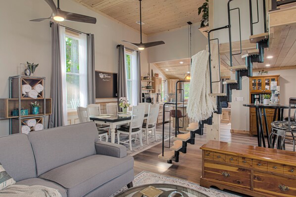 A spacious living area with a sofa, dining table, and chairs, a staircase with a railing, and a kitchen area visible in the background.