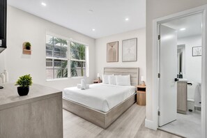 Ein modernes Hotelzimmer mit einem großen Bett, einem Nachttisch, einem Fenster mit Blick und einem durchgegangene Tür erkennbaren Badezimmer.