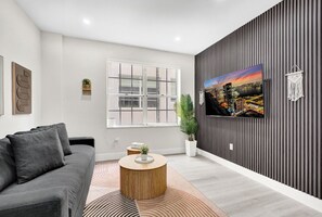 Ein modernes Wohnzimmer mit einem grauen Sofa, einem runden Couchtisch, einem großen Fenster, einem Stadtbildgemälde und einer Topfpflanze.