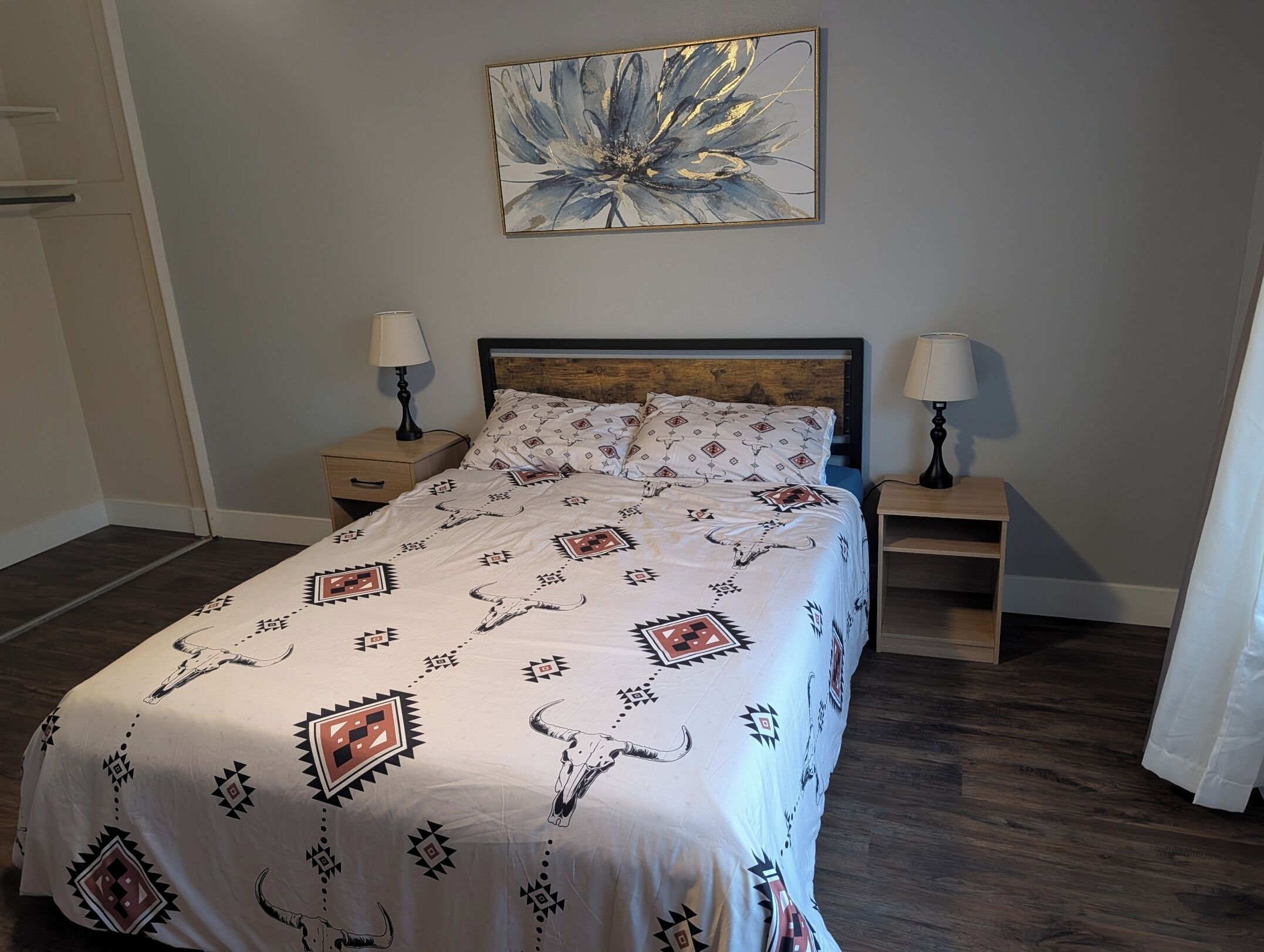A bedroom with a bed, two bedside tables with lamps, a wooden headboard, a wall-mounted abstract painting, and a patterned bedspread.