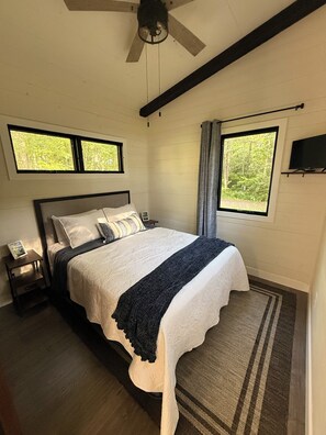 Un dormitorio con una cama, dos mesitas de noche, un ventilador de techo, dos ventanas con cortinas y un televisor en la pared.
