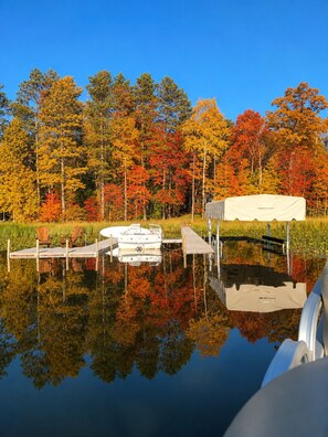Un paisaje tranquilo junto al lago, con un muelle, un bote cubierto y una hilera de árboles con follaje otoñal.