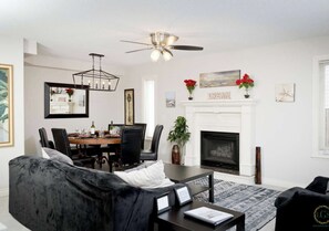 A living room with a fireplace, a dining area, and a ceiling fan. - Arbour Crest by Grenadier Luxury suites (Calgary)