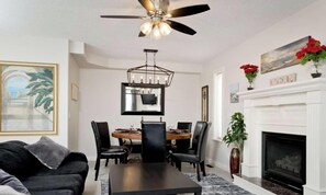 A living room with a ceiling fan, a fireplace, a dining table with chairs, and a sofa. - Arbour Crest by Grenadier Luxury suites (Calgary)