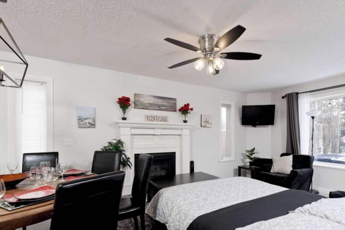 A hotel room with a bed, a dining table with chairs, a fireplace, a ceiling fan, a TV, and a window with a view of trees.