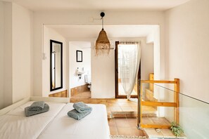 Une chambre d’hôtel comprenant un lit, une chaise en bois, une salle de bain avec un lavabo et un miroir, et un escalier menant à l’étage supérieur.