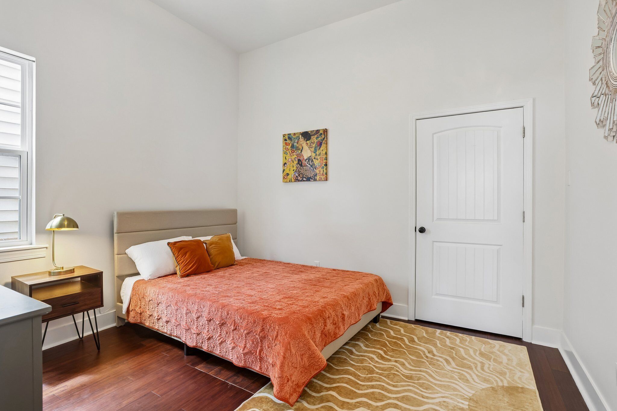 Um quarto com cama, mesa de cabeceira, luminária, porta e uma obra de arte pendurada na parede.