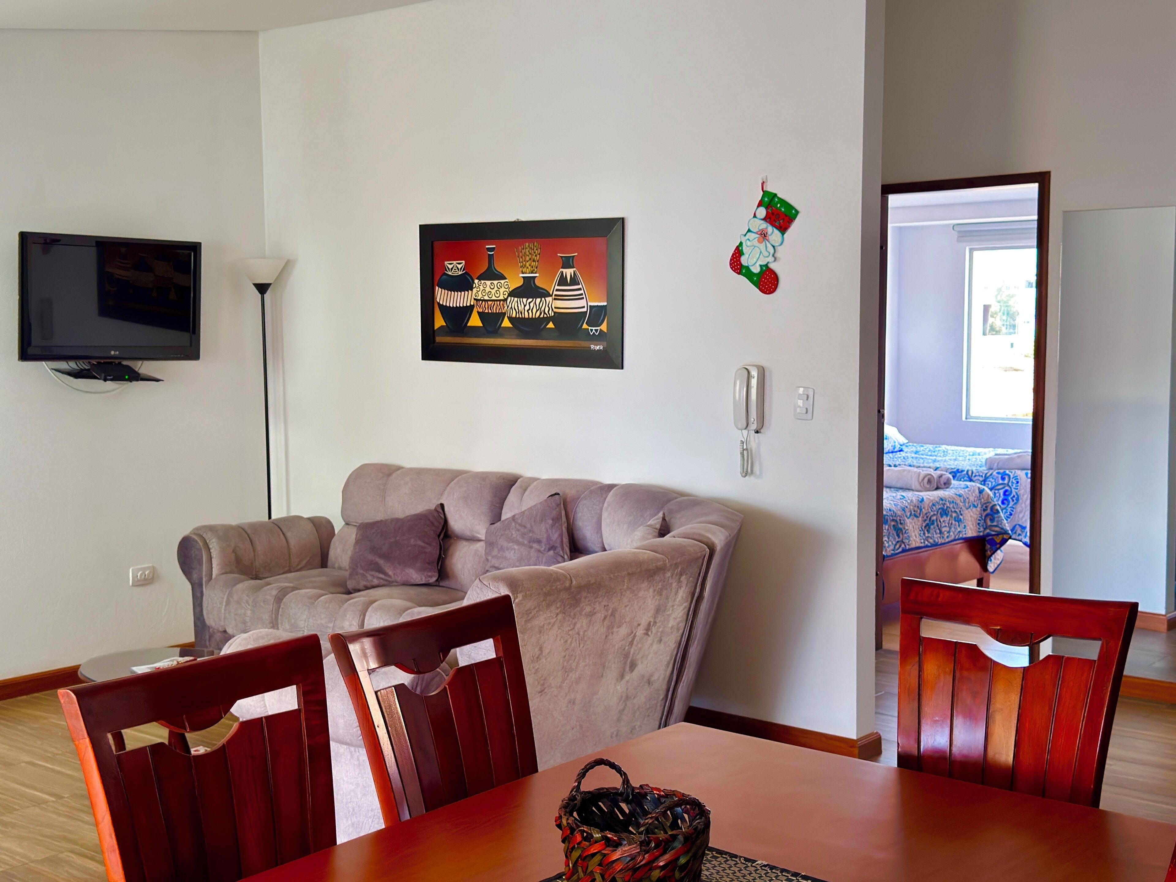 Een woonkamer met een bank, stoelen, een televisie, een ingelijste afbeelding, een telefoon en een deur die leidt naar een slaapkamer met een bed.