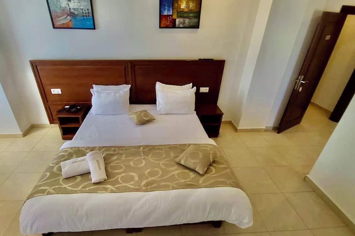 Una habitación de hotel con una cama grande, dos mesitas de noche, una lámpara de noche, un cabecero de madera y dos cuadros en la pared.