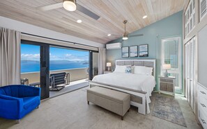 A bedroom with a large bed, a bench, a blue chair, a balcony with a view, and a ceiling fan.