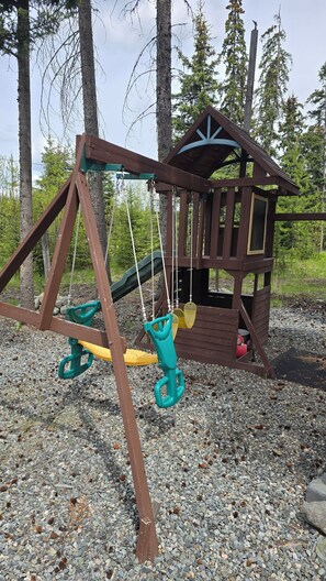 Ein hölzerner Spielplatz mit Schaukel und Rutsche, umgeben von Bäumen und Kies.