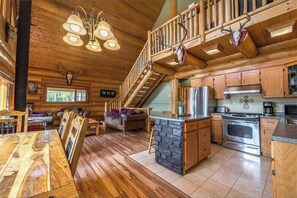 L’intérieur d’une cabane en rondins, avec un escalier en bois, une cuisine dotée d’un îlot central en pierre et un coin repas avec une table en bois.