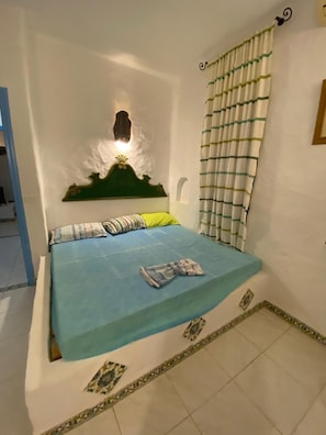A bedroom with a bed, a headboard, a wall-mounted light, a window with curtains, and a tiled floor.