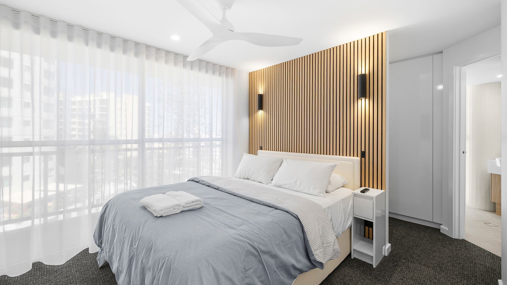 A modern hotel room with a large bed, a ceiling fan, a wooden accent wall, and a view through sheer curtains.