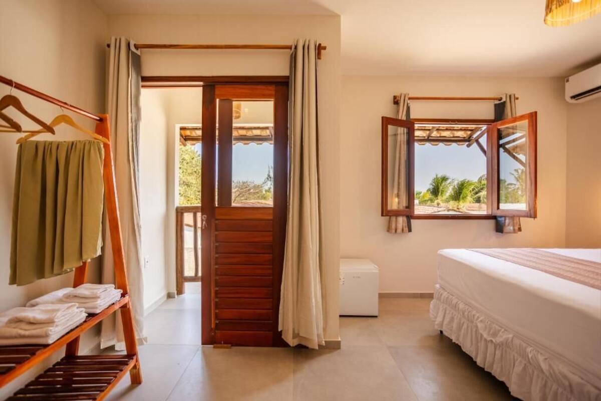 Quarto de hotel com cama, janela com vista, porta de madeira e porta-toalhas.