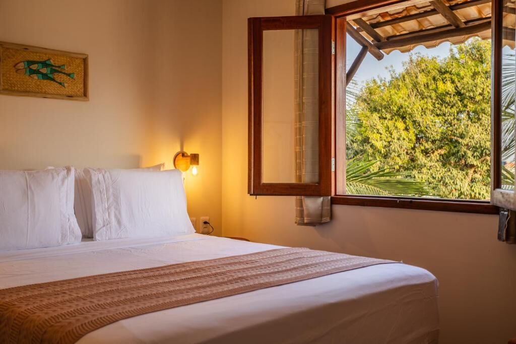 Habitación de hotel con cama, espejo en la pared, ventana con vista a árboles, y lámpara de pared.