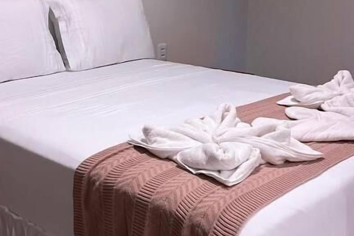 Cama bem arrumada com lençóis brancos, um cobertor marrom e uma toalha sobre a cama.