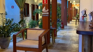 A hotel corridor with wooden pillars, a bench, potted plants, and hanging lights.