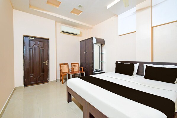 A hotel room with a bed, a desk, two chairs, a wardrobe, and an air conditioning unit.