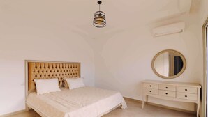 Een slaapkamer met een knopen hoofdeinde, een bed met een witte dekbedovertrek, een nachtkastje met lades, een ronde spiegel en een hanglamp.