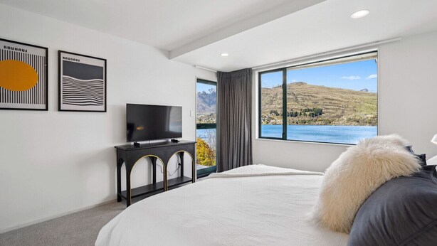 Un dormitorio con cama, televisor sobre un mueble, ventana grande con vistas a un lago y colinas, y dos obras de arte enmarcadas en la pared.