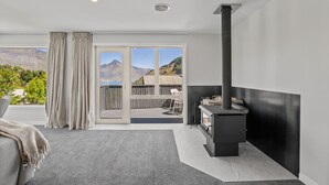 A modern room with a large sliding glass door leading to an outdoor deck, a built-in wood stove, and a view of mountains and a lake. - Alpine Lakeview Residence (Queenstown)