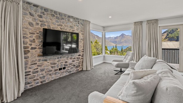 A modern living room with a stone wall, a flat-screen TV, a gray sofa, and a view of mountains. - Alpine Lakeview Residence (Queenstown)