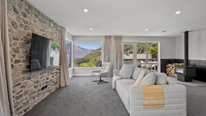A modern living room with a stone wall, a large window with a mountain view, a fireplace, and a cozy seating area. - Alpine Lakeview Residence (Queenstown)