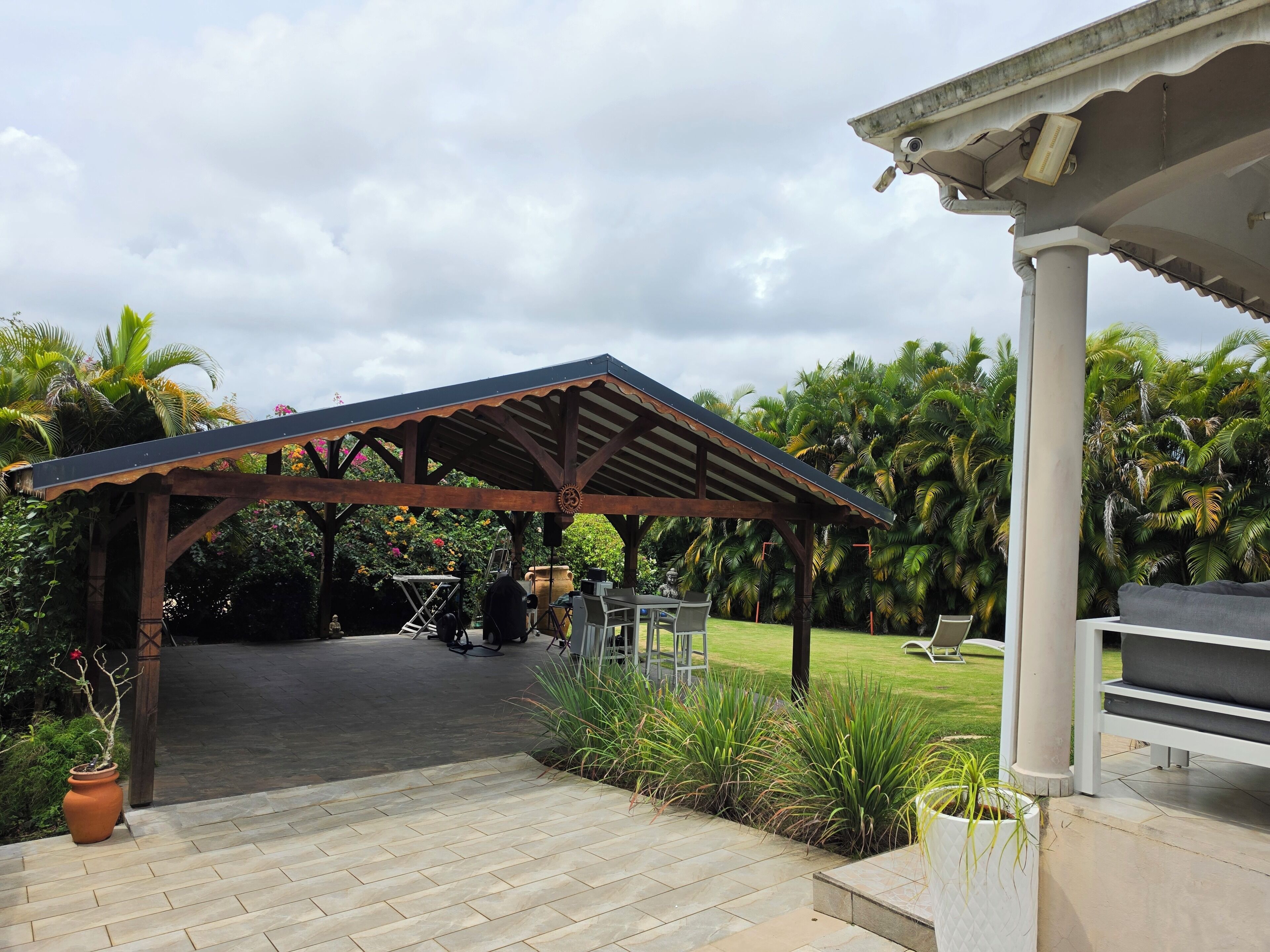 A covered outdoor area with a wooden structure, a grill, and a lawn with palm trees.