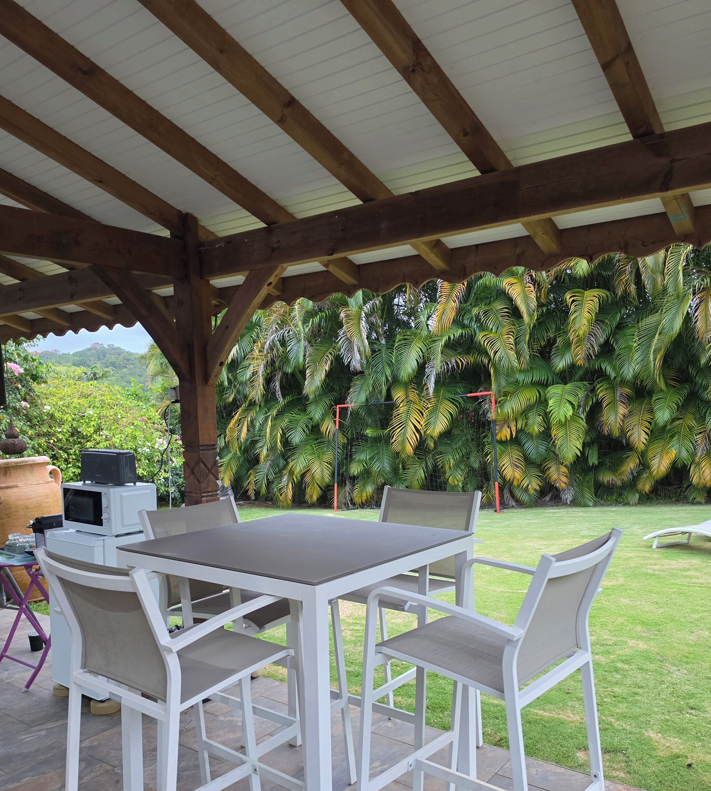 An outdoor patio with a white table and chairs, a covered roof, and a backdrop of lush greenery.