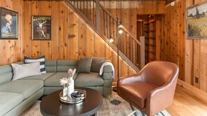 Una habitación con paneles de madera, un sofá verde, un sillón de cuero marrón, una mesa de centro redonda con objetos decorativos y una escalera con barandillas de madera.