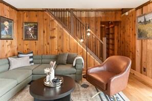 A wooden-paneled room with a green sofa, a brown leather chair, a round coffee table with decorative items, and a staircase with wooden railings.
