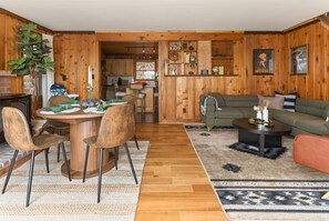 A wooden interior with a dining area, sofa, and a kitchen visible through an open door.