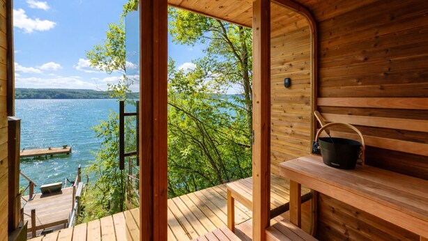 Una cabaña de madera con vista a un lago y árboles, además de una sauna y un muelle.