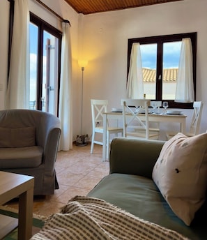A living room with a sofa, a dining table with chairs, a lamp, and a view of the outside through a window.