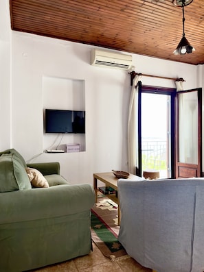 A living room with a green sofa, a television mounted on the wall, a wooden ceiling, and a window with curtains.