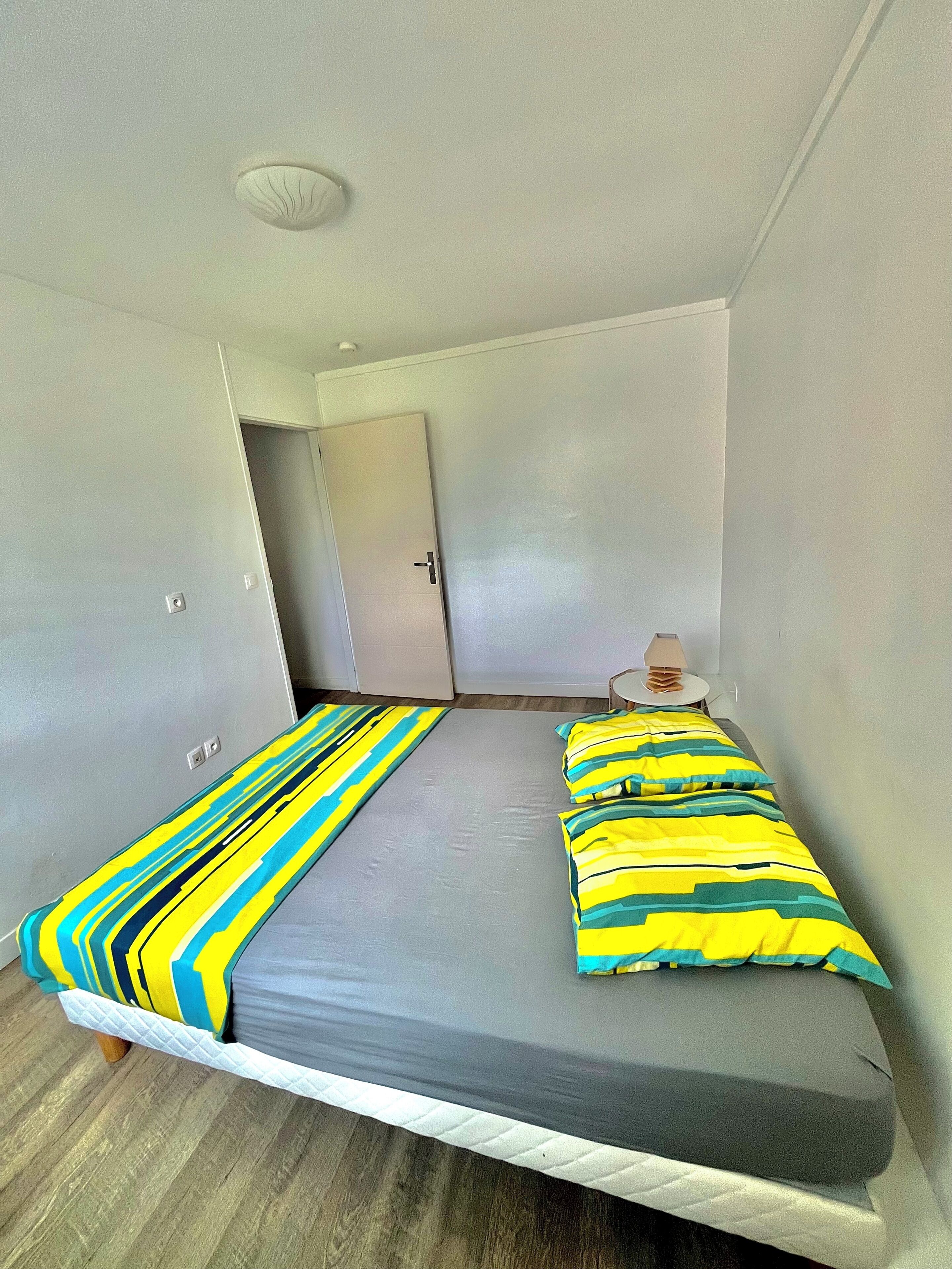 A small, single-bed room with a grey mattress, striped pillows, a wooden floor, a white door, and a ceiling light fixture.