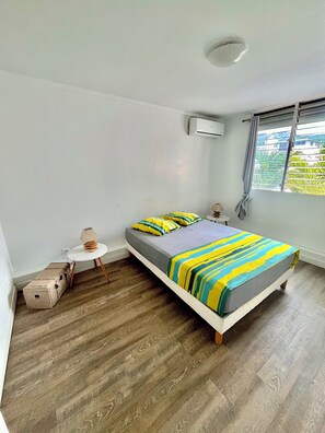 A bedroom with a bed, a small table, a lamp, a suitcase, and an air conditioning unit. - Le Balcon Marina - 3 rooms sea view (Pointe-à-Pitre)