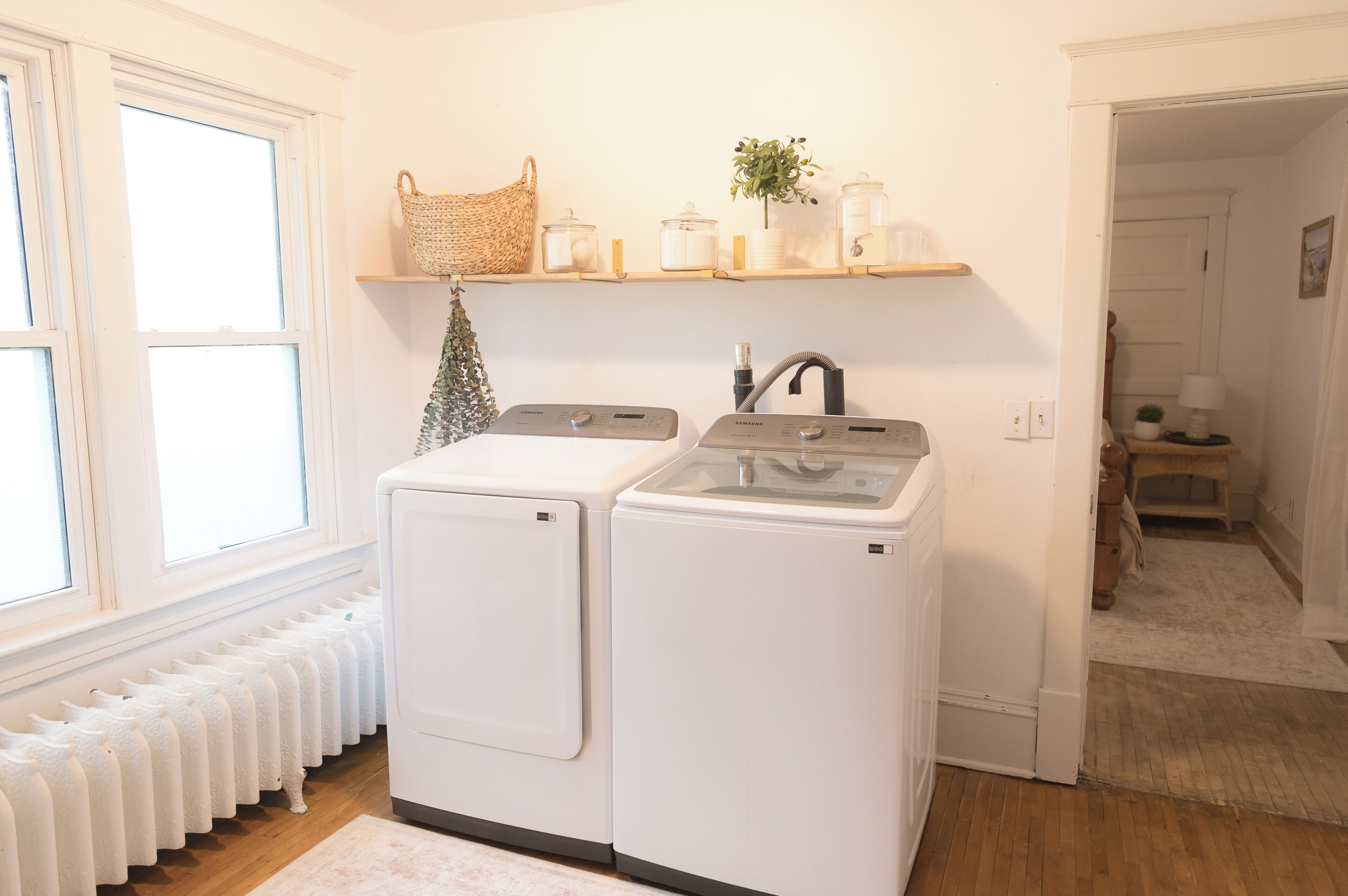 A laundry room with two white dryers, a radiator, a shelf with jars and a plant, and a wicker basket.